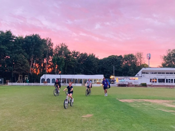 Cyclists crossing the cricket ground beneath a pink sunset